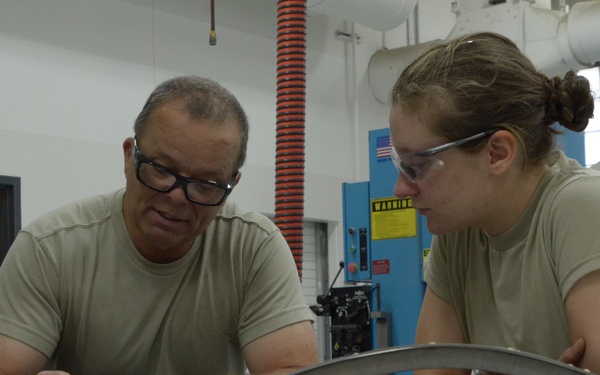 Two individuals wearing olive/tan military-style t-shirts are working together at a workbench in an industrial or military maintenance shop. The person on the left is writing on a document while both examine a curved metal component placed on the surface. The setting appears to be a fabrication or metalworking facility.