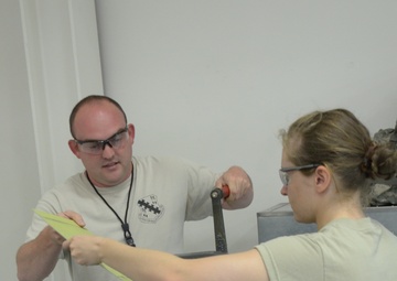 Two individuals in U.S. Air Force uniforms (ABU - Airman Battle Uniform) are working together at what appears to be a sheet metal brake or bending machine in a workshop or training facility. One person, a male wearing a tan t-shirt with a military unit insignia and safety glasses, is guiding a yellow material strip, while the female airman in full ABU uniform operates the machine's handle. Both are wearing protective eyewear, indicating adherence to safety protocols.