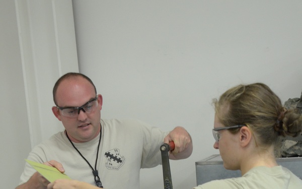 Two individuals in U.S. Air Force uniforms (ABU - Airman Battle Uniform) are working together at what appears to be a sheet metal brake or bending machine in a workshop or training facility. One person, a male wearing a tan t-shirt with a military unit insignia and safety glasses, is guiding a yellow material strip, while the female airman in full ABU uniform operates the machine's handle. Both are wearing protective eyewear, indicating adherence to safety protocols.