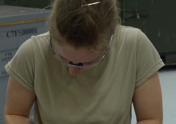 A woman wearing safety glasses and a tan/khaki t-shirt is working at a workbench, marking or scribing lines on a metal sheet using a tool. She appears to be in an industrial or military maintenance facility, with equipment and labeled containers visible in the background. The work surface shows signs of use, and a template or drawing appears to be placed on top of the metal sheet.