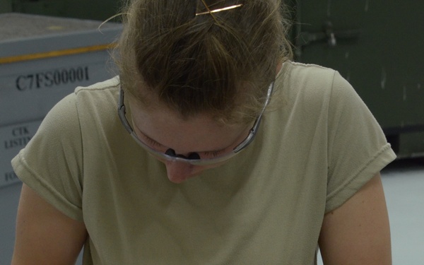 A woman wearing safety glasses and a tan/khaki t-shirt is working at a workbench, marking or scribing lines on a metal sheet using a tool. She appears to be in an industrial or military maintenance facility, with equipment and labeled containers visible in the background. The work surface shows signs of use, and a template or drawing appears to be placed on top of the metal sheet.
