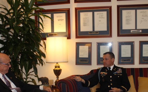 Two individuals are seated in a formal office setting engaged in conversation. One person is wearing a U.S. Army dress uniform with visible rank insignia and service ribbons, while the other is dressed in civilian business attire. The room appears to be an official office decorated with numerous framed documents or certificates on the wall.