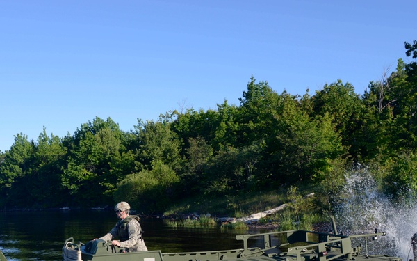 A military engineer operates an olive-drab assault/utility boat on a calm river or lake, while a large military bridging or engineering vehicle on the right bank appears to be launching or deploying equipment into the water. The scene suggests a military river-crossing or bridging operation exercise. The setting is a wooded shoreline under clear blue skies.
