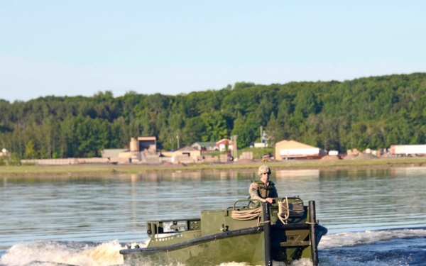 A military personnel in combat uniform and helmet operates an olive-drab military watercraft at speed across a calm river or lake. The vessel appears to be a military assault or engineer boat, creating a visible wake. Buildings and dense green treeline are visible on the far shoreline in the background.