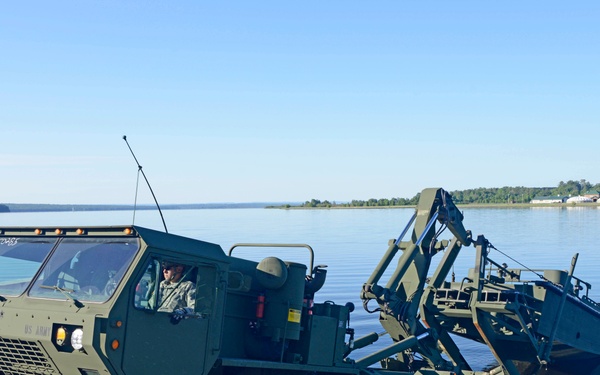 A US Army military vehicle, specifically an Improved Ribbon Bridge (IRB) carrier truck, is being driven into a body of water from a boat ramp or launch area. The large 8-wheeled tactical truck is towing or deploying bridge sections into the water, likely during a military engineering training exercise. The setting appears to be a calm lake or river with forested shoreline visible in the background.