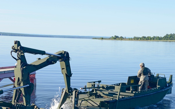 A military amphibious bridging operation is underway on a calm body of water. A soldier in military uniform operates what appears to be an Improved Ribbon Bridge (IRB) or similar military bridging system, with a crane/articulating arm mechanism actively deploying or connecting bridge sections. The equipment is olive drab green, consistent with U.S. Army engineering assets.