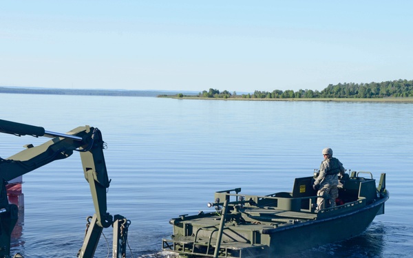 A military engineer soldier operates an olive-drab colored watercraft, likely an Improved Ribbon Bridge (IRB) boat or similar military bridging vessel, being launched or retrieved from a body of calm water. A large military crane or handling mechanism on a trailer is visible on the left, likely used to deploy or recover the watercraft. The setting appears to be a lake or wide river with forested shoreline in the background under clear skies.