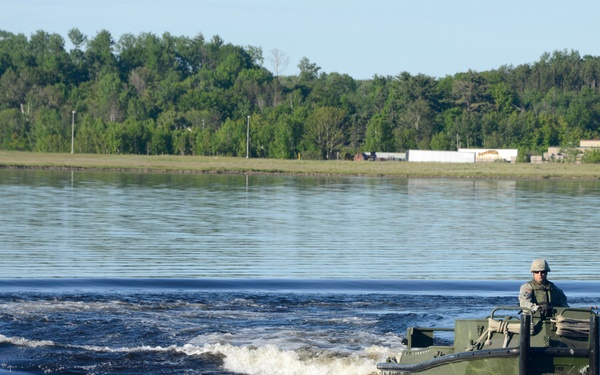 A military soldier is operating an olive-drab amphibious assault craft on a calm body of water, creating a visible wake behind the vessel. The setting appears to be a training exercise at a military installation, with a flat shoreline, tree line, and what appears to be a warehouse or facility structure visible in the background. The image was taken during daylight under clear sky conditions.