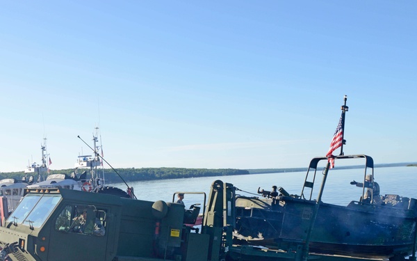 A large military tactical truck, appearing to be an LMTV or HEMTT variant in olive drab green, is being driven off or onto a river landing ramp near a waterway. A soldier in ACU camouflage uniform is directing the vehicle from the shoreline. An American flag is visible mounted on the rear of the truck, and military watercraft are docked in the background.