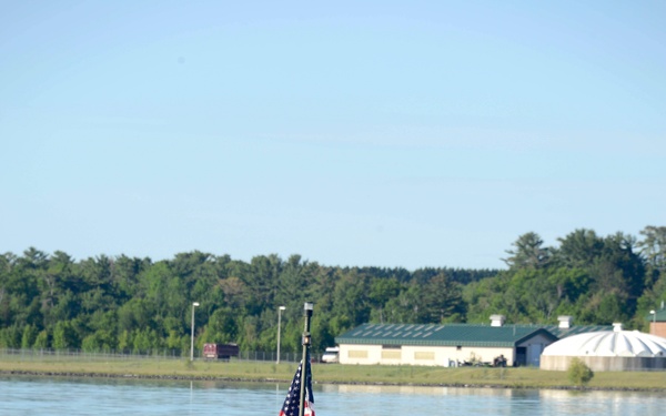 A uniformed military soldier operates a small camouflage-painted military watercraft on a calm river or lake. An American flag is mounted on a pole at the rear of the vessel. Buildings and tree lines are visible on the far shore in the background.