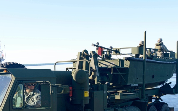 A US Army heavy tactical truck (HEMTT or LMTV-class vehicle) is transporting a military patrol boat toward a water launch ramp. Several soldiers in ACU (Army Combat Uniform) are visible, with one directing the vehicle from outside and others positioned on the boat. The scene appears to be a boat launch exercise or operation at a waterfront facility.