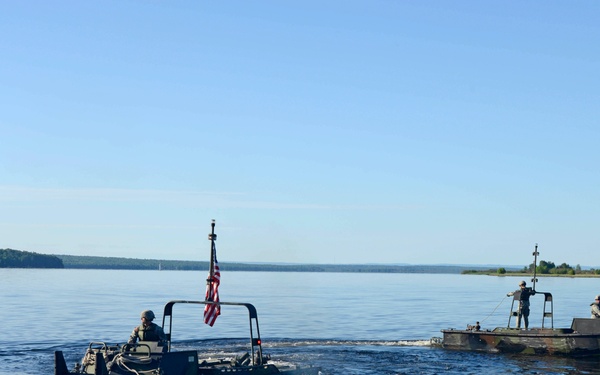 Two military watercraft are operating on a calm, wide body of water near a dock with red metal framing. The nearer vessel displays an American flag and is operated by a soldier in military gear, while a second larger flat-deck military craft with multiple personnel is visible in the background. The scene appears to depict a military training or operational exercise on an inland lake or river.