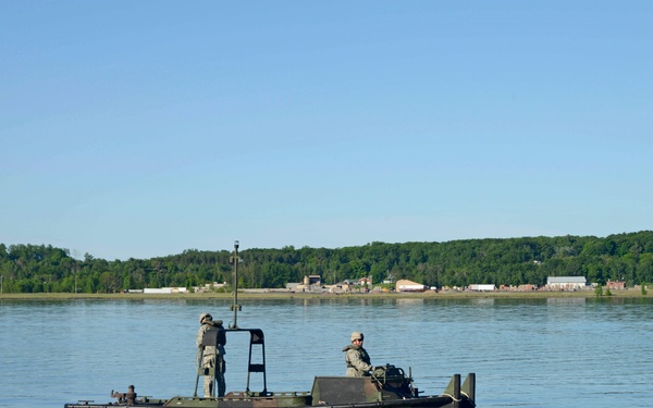 A military utility boat painted in olive drab/camouflage colors is underway on a calm body of water. Two personnel in military uniforms are aboard — one seated at the helm and one standing near the bow. A developed shoreline with buildings and dense tree line is visible in the background.
