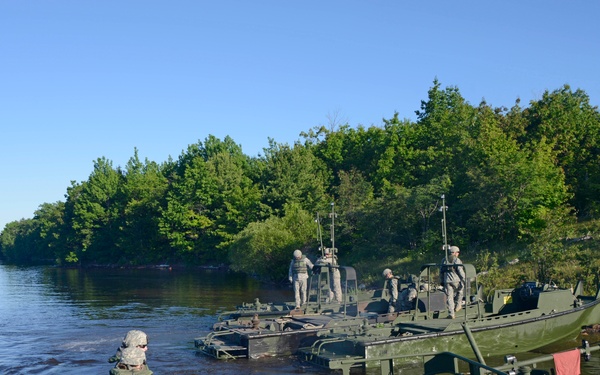 Military personnel in camouflage uniforms are operating olive-drab colored engineer boats along a riverbank surrounded by dense green trees. The scene appears to depict a military bridging or river-crossing operation, with multiple soldiers working on and around the vessels. A structure or building is faintly visible in the background across the river.