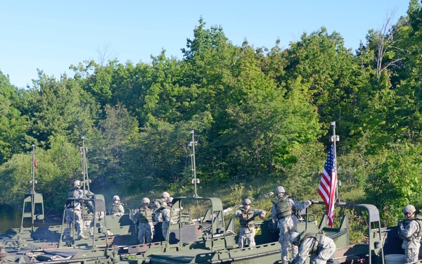Three military assault/engineer boats are moored together along a riverbank or lakeshore, with multiple soldiers in camouflage uniforms aboard each vessel. An American flag is prominently displayed on the rightmost boat. The scene appears to be a military training exercise or operational staging event in a wooded, inland waterway environment.