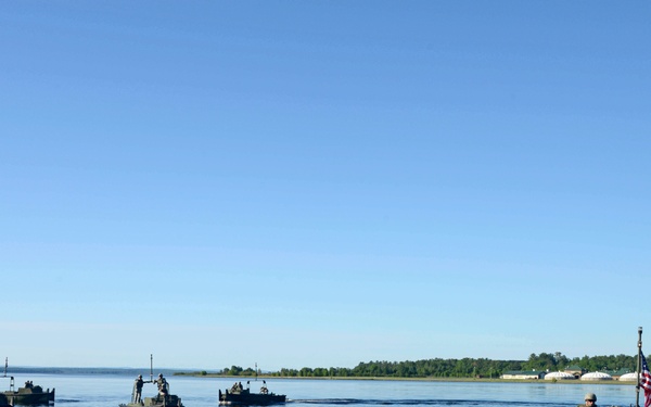 A formation of military watercraft carrying uniformed personnel is operating on a calm lake or river, viewed from a dock. The nearest vessel displays a US flag on a pole, and multiple boats are arranged in a loose formation moving away from the dock. The setting appears to be a military training exercise conducted in a forested inland waterway environment.