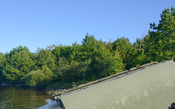 A military amphibious bridging or ferry section is being launched into a body of water from a wheeled military vehicle. The olive-drab colored pontoon/float section is tilted at an angle as it enters the water, creating a splash. The operation appears to be taking place at a riverbank or lake shore with dense green tree line in the background.