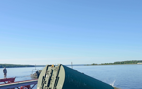 Military personnel are launching a large olive-drab pontoon or bridge section into a wide river or lake from a heavy military truck/transporter. The equipment appears to be part of a tactical bridging or floating bridge system, likely an Improved Ribbon Bridge (IRB) or similar military bridging asset. The operation is taking place on a calm waterway with forested shoreline visible in the background.
