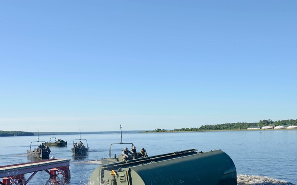 A military bridging or amphibious engineering operation is underway on a wide river or lake. A large green military float bridge section or amphibious vehicle is being launched into the water from a ramp, with personnel visible on top. Additional military watercraft and support boats are visible in the background.