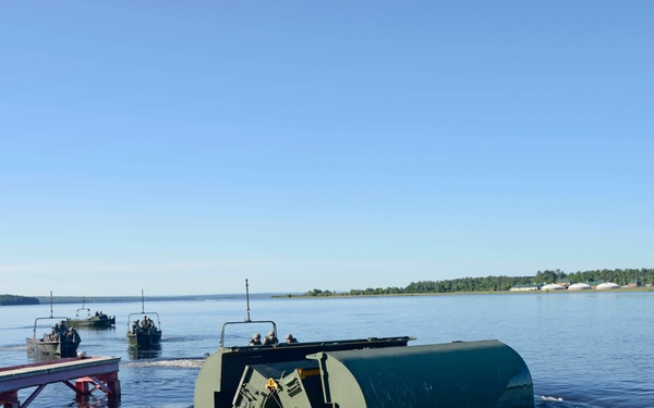 A military bridging or river-crossing operation is underway on a wide river or lake. A large green military pontoon/float unit is being launched from a ramp into the water, while additional military watercraft are visible operating in the background. Personnel in military uniforms are present on the dock and aboard the vessels.