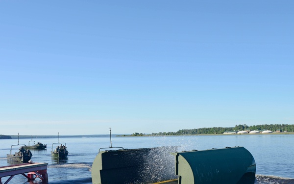 A military bridging or floating pontoon system is being launched into a wide body of water from a ramp or dock. The green military equipment appears to be entering the water at an angle, with water splashing around it. Additional military watercraft are visible in the background, suggesting a coordinated military engineering exercise.
