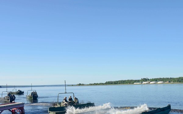 Military bridging or amphibious engineering vehicles are being launched into a wide river or lake from a shoreline ramp. Multiple olive-drab colored military watercraft, likely Improved Ribbon Bridge (IRB) or similar pontoon bridge sections, are entering the water with personnel aboard. The operation appears to be a training exercise conducted in calm, clear weather conditions.