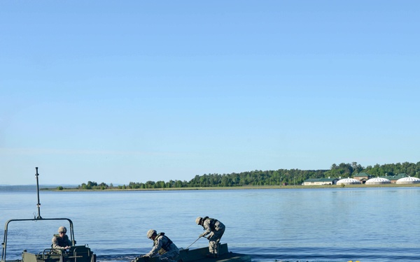 Military personnel are conducting a bridging or river-crossing operation on a calm body of water. Soldiers in camouflage uniforms are operating green military pontoon bridge sections and an assault boat. A military installation with dome-shaped structures is visible on the far shore.