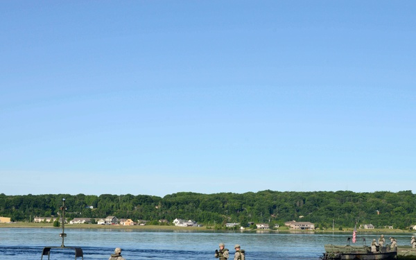 Military personnel are operating pontoon bridge or ferry sections on a wide river during what appears to be a training exercise. Multiple green military watercraft are visible, with soldiers in camouflage uniforms standing on the platforms. A residential area with trees is visible on the far bank.