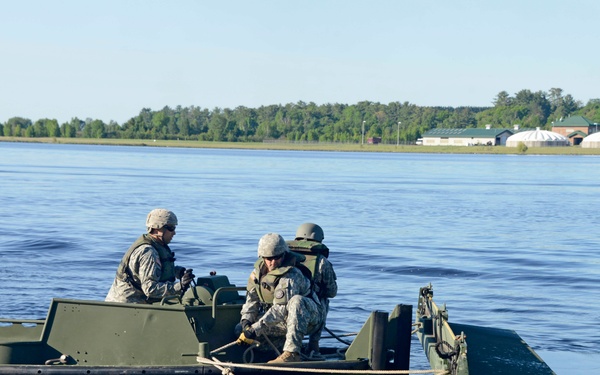 Two military personnel in combat uniforms and helmets are operating a military assault boat on a wide river or lake. They appear to be conducting a bridging or river-crossing operation, as evidenced by a rope connecting their boat to a floating bridge/raft section visible on the right. Buildings and tree lines are visible on the far shore in the background.