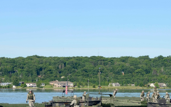 Military personnel are operating green amphibious bridging/ferry vessels on a wide river or lake during what appears to be a training exercise. Multiple soldiers in combat uniforms and life vests are visible aboard at least two separate watercraft. A residential area with trees and an American flag is visible on the far shore.