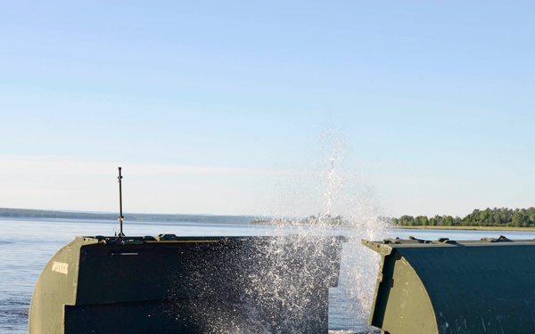 A military amphibious bridging or ferry unit is being launched into a wide body of water, with water splashing dramatically as it enters. The olive-drab colored vessel appears to be a pontoon bridge section or ribbon bridge component unfolding upon water entry. A second military watercraft is visible in the background, and a dock structure is partially visible on the left.