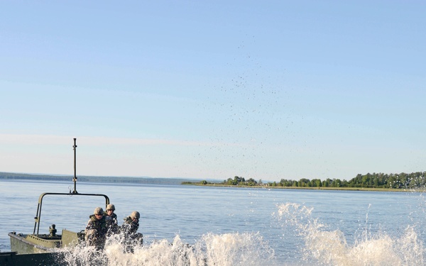 A military pontoon or assault bridge section is being launched or maneuvered on a wide river or lake, generating large water splashes. Several soldiers wearing helmets are visible operating the vessel. A second military watercraft is visible in the background to the left.