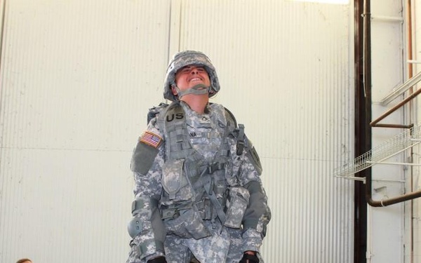 A U.S. Army soldier in full combat gear including ACU (Army Combat Uniform), helmet, body armor, and knee pads is standing elevated on a platform or equipment, looking upward. Another soldier in ACU is crouching nearby observing, while two civilians in dark maroon shirts stand in the background, one taking notes. The setting appears to be an indoor training or testing facility, possibly related to parachute or equipment fitting evaluation.