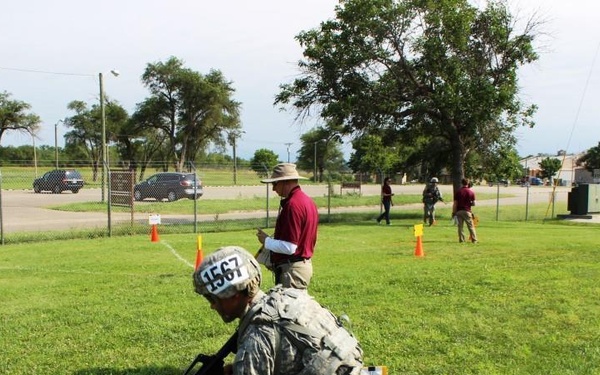 A soldier wearing full combat gear and a helmet marked with the number '1567' is running in a low crouch while carrying a rifle, appearing to participate in a military training exercise or competition. The course is marked with orange traffic cones on a grassy field, with a civilian observer in a red shirt and hat watching nearby. Additional personnel and vehicles are visible in the background near a fenced area.