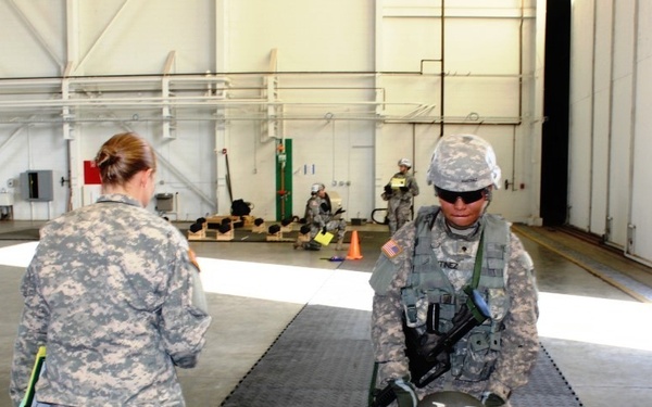 The image shows U.S. Army soldiers in ACU (Army Combat Uniform) inside what appears to be a large hangar or training facility. A soldier with the name tape 'TINEZ' (partially visible, likely 'MARTINEZ') is working with a training mannequin strapped to a seat, while another soldier holding a clipboard or folder observes. Additional soldiers are visible in the background engaged in similar training activities.