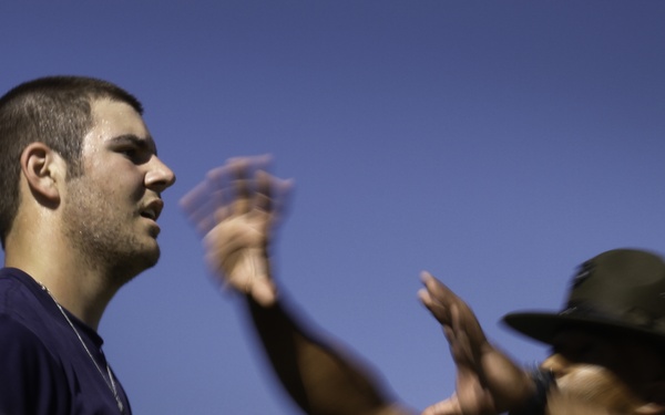 Two individuals are shown outdoors against a clear blue sky. One person wearing a navy blue t-shirt with a name tag patch stands at attention, while a second person in military camouflage uniform and a drill instructor-style campaign cover (hat) gestures animatedly with both hands in motion blur. The scene appears to depict a military training or recruitment scenario, likely involving a drill instructor addressing a recruit.