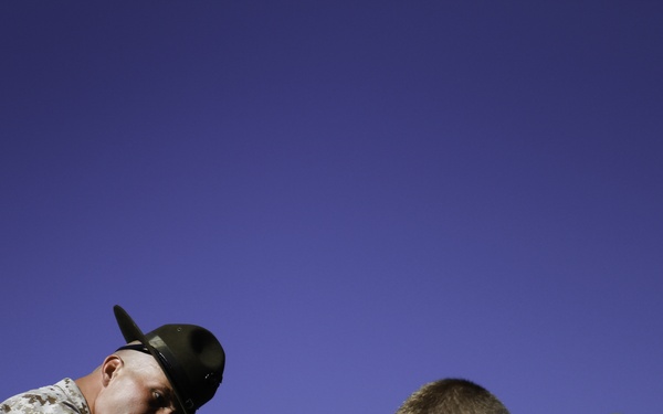 A military drill instructor wearing MARPAT camouflage uniform and a campaign cover (drill sergeant hat) is leaning forward and pointing at a recruit dressed in an olive green t-shirt. The recruit is bent over, appearing to be in a submissive or exhausted posture. The image is shot from a low angle against a clear blue sky, emphasizing the confrontational dynamic between the two individuals.