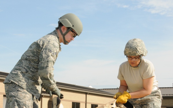 Two military personnel in camouflage uniforms and helmets are working together outdoors to set up or break down a large tan/beige military tent or shelter on a grass field. Both individuals are wearing protective gloves and appear to be on a military installation based on the brick buildings visible in the background. The activity appears to be a field training exercise or base operations task.
