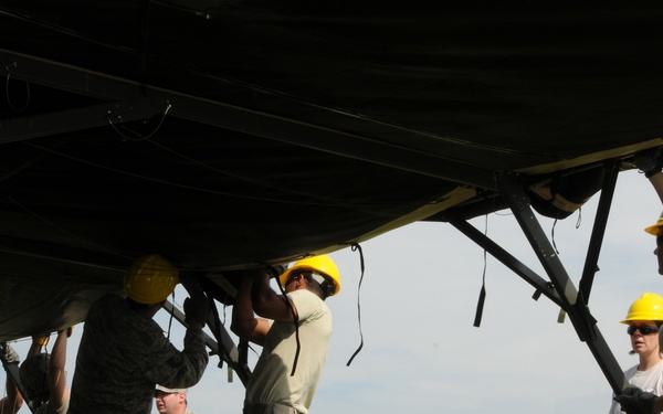 A group of military personnel in U.S. Air Force ACU/ABU camouflage uniforms are collectively assembling or raising a large military tent structure on a grass field. Several individuals are wearing yellow hard hats, indicating a construction or setup safety protocol. The activity appears to be taking place on or near a military installation, with a brick building visible in the background.