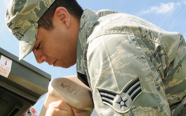 A U.S. Air Force Senior Airman in ABU (Airman Battle Uniform) is performing a maintenance or technical task on military equipment, leaning over and working with a tool or component. A second service member is visible in the background, partially obscured. The image is taken from a low angle, emphasizing the rank insignia on the primary subject's sleeve.