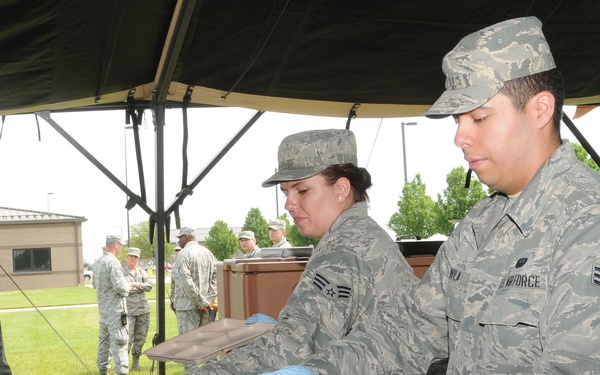 U.S. Air Force personnel are participating in an outdoor field dining event under a military canopy tent. Service members in Airman Battle Uniforms (ABU) are being served food from insulated food transport containers by fellow airmen wearing blue food-service gloves. The setting appears to be a base or training area with additional personnel visible in the background.