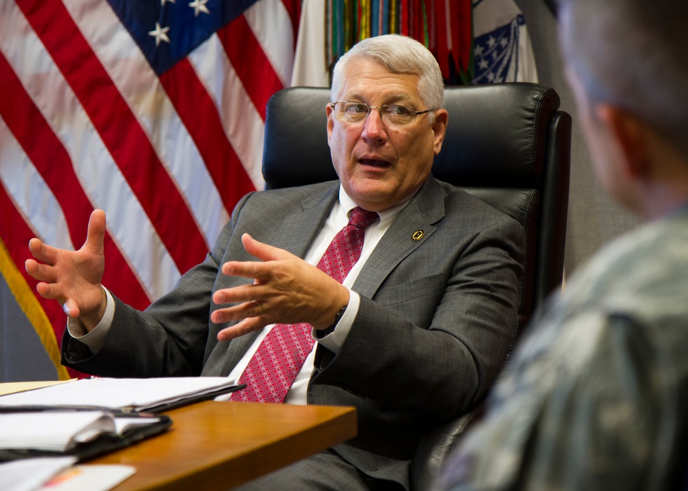 A senior civilian official in a gray suit and red tie is seated at a desk in what appears to be a formal government or military office, engaged in an animated discussion with gesturing hands. Multiple flags are visible in the background, including the United States flag and what appear to be military command or unit flags. A person in military camouflage uniform is partially visible in the foreground.