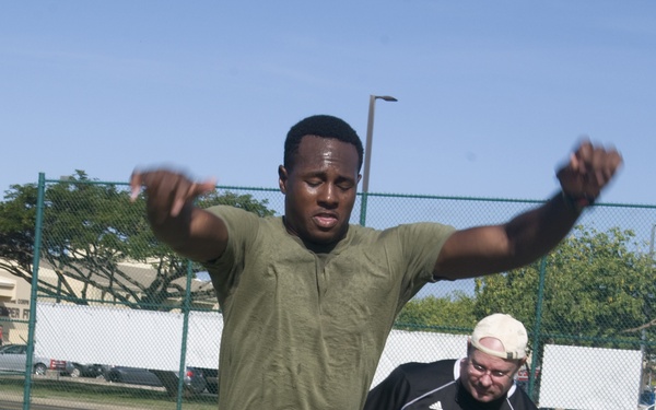 A man wearing a military-style olive green t-shirt and desert camouflage trousers is performing a standing broad jump or long jump on a grass field. A second man in a black Adidas shirt and backwards cap observes from behind, likely acting as a judge or trainer. Training equipment including a green measuring device, an orange cone, and a water bottle are visible on the ground.