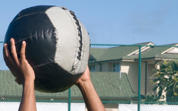 A man wearing an olive green military-style t-shirt is lifting a large medicine ball overhead with both arms extended on a grassy outdoor field. He appears to be engaged in a physical fitness training exercise, with his eyes closed and visible perspiration on his face. A second person in military attire is partially visible on the left edge of the frame.