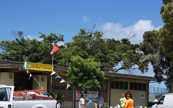 A commercial parking lot scene in front of a building labeled 'K-Bay's Garden' with the address 1255 visible. Two white vehicles branded 'NARITO Sheet Metal &amp; Mechanical' — a pickup truck and a cargo van — are parked in the lot. Several workers are standing and conversing in the parking area, which is decorated with colorful bunting flags.