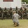 A military training or demonstration session is taking place outdoors at what appears to be a military installation. A soldier in US Army ACU (Army Combat Uniform) pattern camouflage is kneeling on the ground facing a group of approximately eight soldiers seated on steps, who appear to be wearing Jamaican Defence Force uniforms with distinctive patches. The scene suggests a joint military training exercise or instruction session between US and Caribbean military personnel.