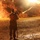 A person in military-style camouflage uniform is filming a nighttime scene with a professional video camera mounted on a monopod. Large fires are burning on both sides of an earthen berm or range structure, illuminating the scene with intense orange light. The setting appears to be a military training range or controlled demolition/fire exercise.