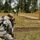 A military training exercise is taking place at an outdoor shooting range surrounded by pine trees. A female soldier in US Army ACU camouflage is kneeling in the foreground filming with a video camera, while in the background another soldier in camouflage aims a rifle downrange alongside a civilian in a white shirt. Shooting targets on wooden stands are visible to the right side of the range.