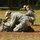 Two military personnel in US Army ACU (Army Combat Uniform) digital camouflage are positioned low to the ground in an outdoor field setting. The soldier in the foreground is operating a Panasonic professional video camera from a kneeling/prone position, while the soldier in the background is using a DSLR camera with a telephoto lens. Both appear to be military public affairs or combat camera personnel documenting an event or exercise.