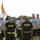 A group of U.S. Army personnel in physical training (PT) uniforms are assembled on a grass field. Two soldiers in the foreground wear black Army PT shirts and shorts with yellow reflective belts, with one holding a unit guidon flag. Behind them, additional soldiers in gray Army PT shirts stand in formation.